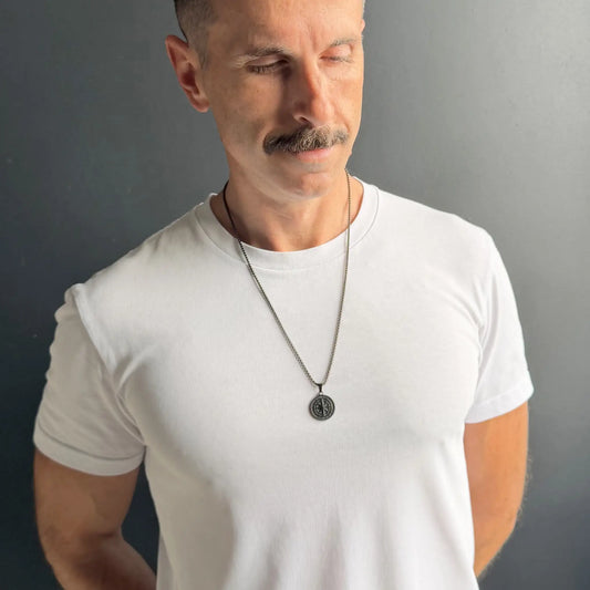 Man with moustache wearing a black compass pendant necklace over a white tshirt against a dark grey background