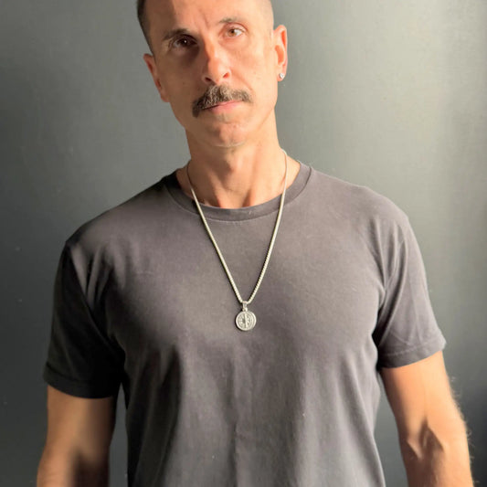 Man with moustache wearing a silver compass pendant necklace over a black tshirt against a dark grey background