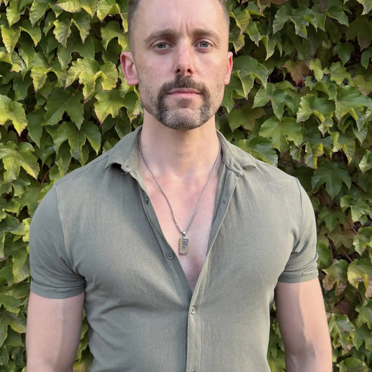 Gold and silver stainless steel dog tag pendant necklace worn by a bearded man with blue eyes with a casual green button up shirt against a healthy hedge