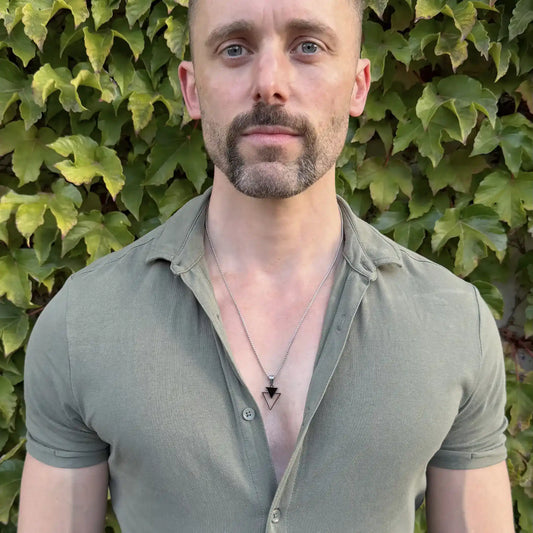 Bearded man standing against a green hedge wearing a stainless steel pendant necklace with a small black triangle stacked over an open triangle and a casual green button shirt