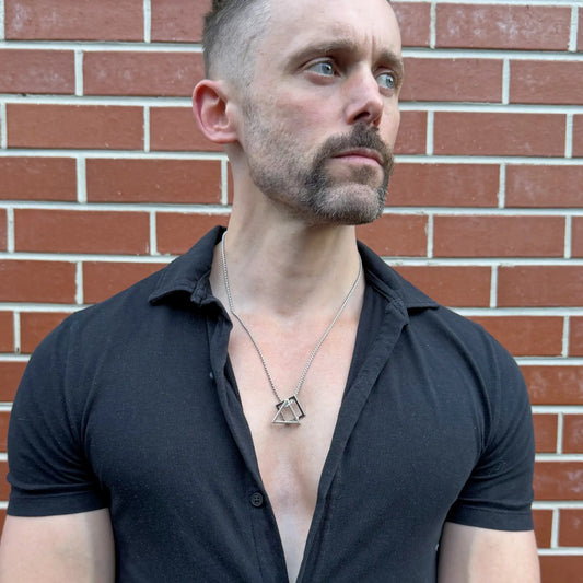 Stainless steel pendant necklace with interlocking square and triangle worn by a bearded man with a black button shirt against a terracotta brick wall