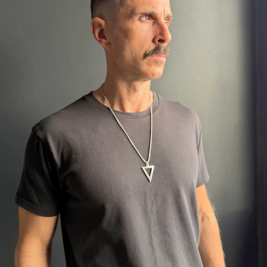 Man with moustache wearing a stainless steel silver triangle pendant necklace over a black tshirt against a dark grey background