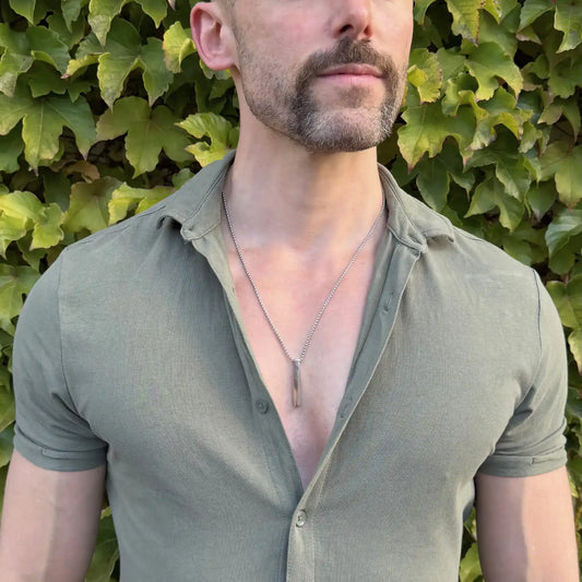 Stainless steel bar pendant necklace worn by a bearded man with a green button shirt against a green hedge
