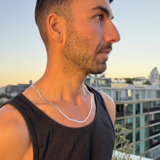Man in black singlet wearing a silver 3mm snake chain necklace outdoors at golden hour, looking sideways with city buildings behind