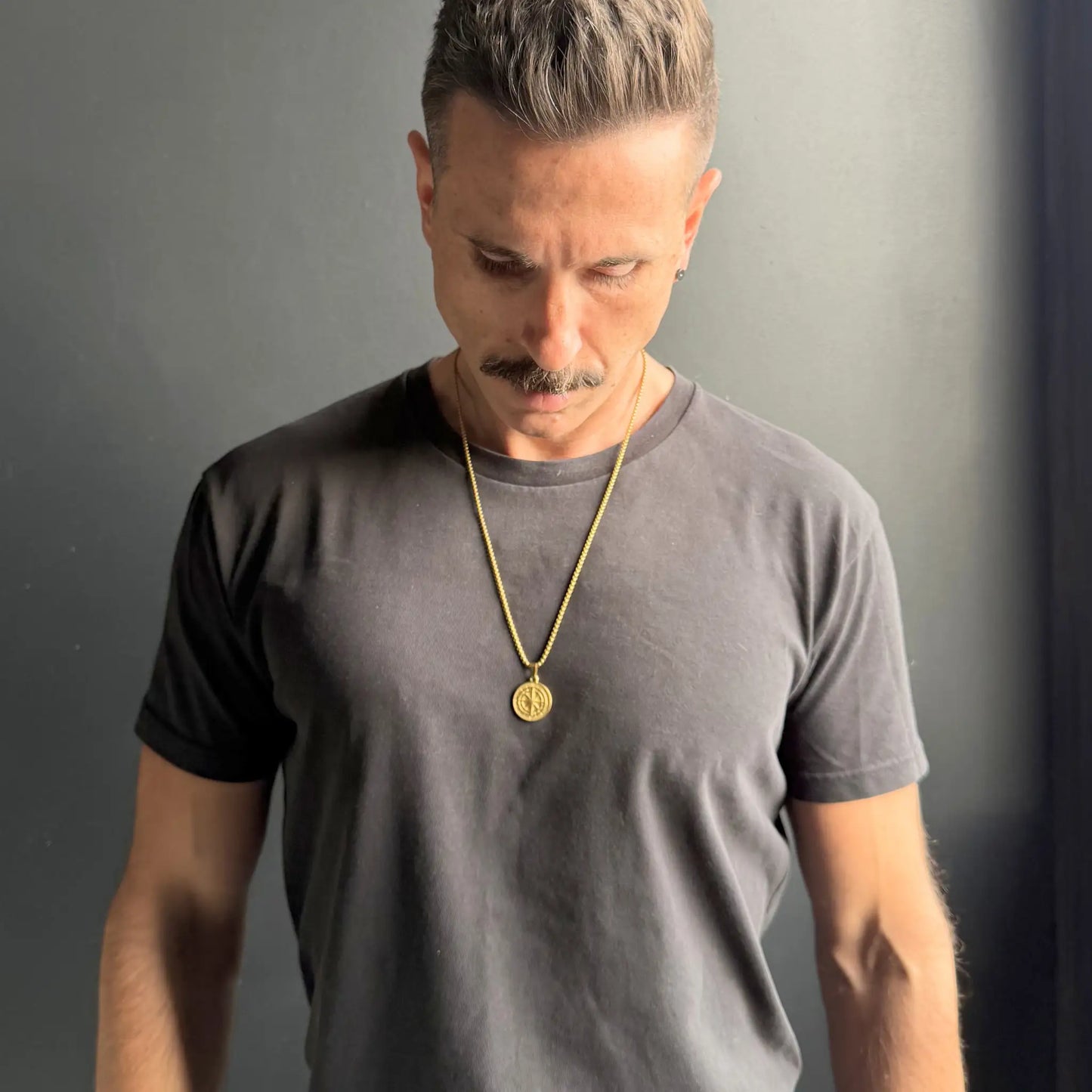 Man with dark hair and moustache wearing a gold compass pendant necklace over a black tshirt against a dark grey background