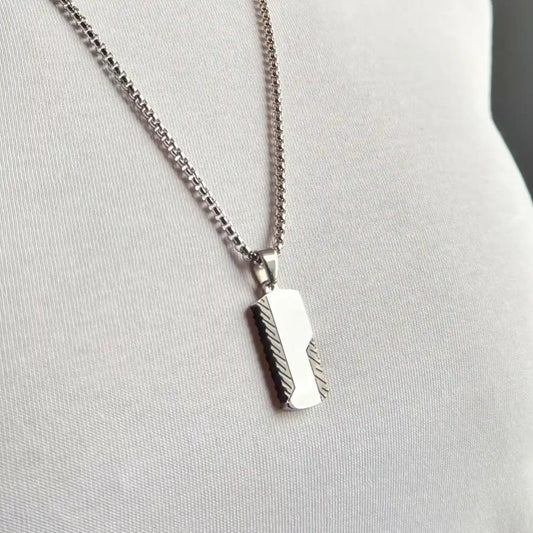 Close up of a silver stainless steel dog tag pendant necklace with black detailing on edges worn against a white tshirt and a dark grey background
