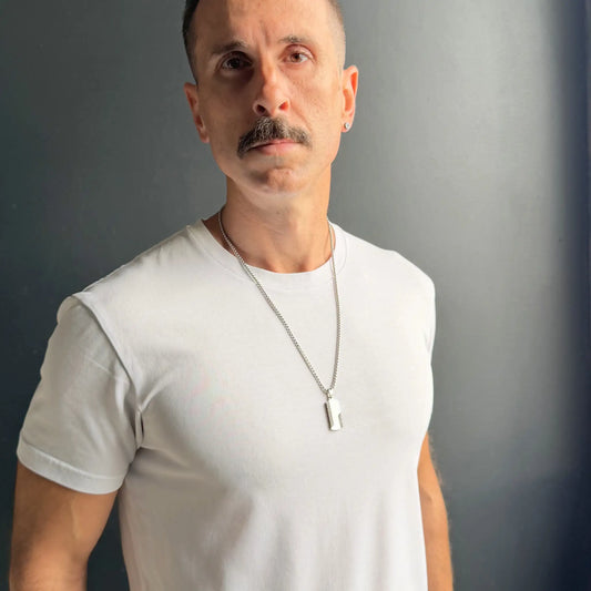 Man with moustache wearing a silver dog tag pendant necklace with black detailing over a white shirt and a dark grey background
