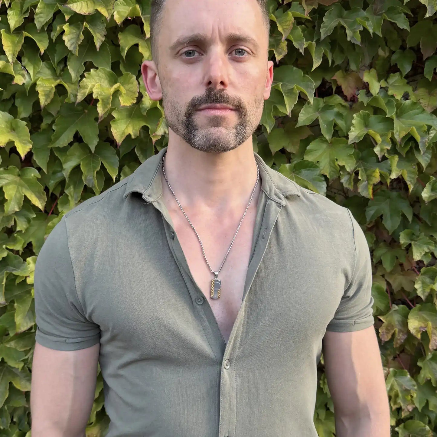 Gold and silver stainless steel dog tag pendant necklace worn by a bearded man with blue eyes with a casual green button up shirt against a healthy hedge