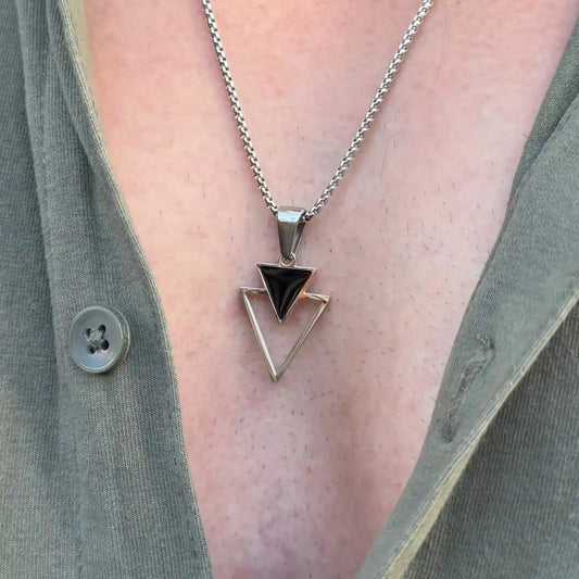 Close up of a stainless steel pendant necklace with a small black triangle stacked over an open triangle worn by a man with a casual green button shirt