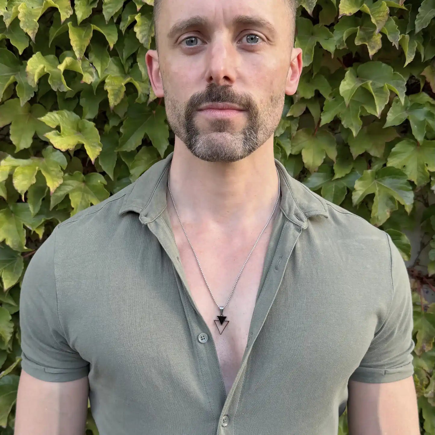 Bearded man standing against a green hedge wearing a stainless steel pendant necklace with a small black triangle stacked over an open triangle and a casual green button shirt