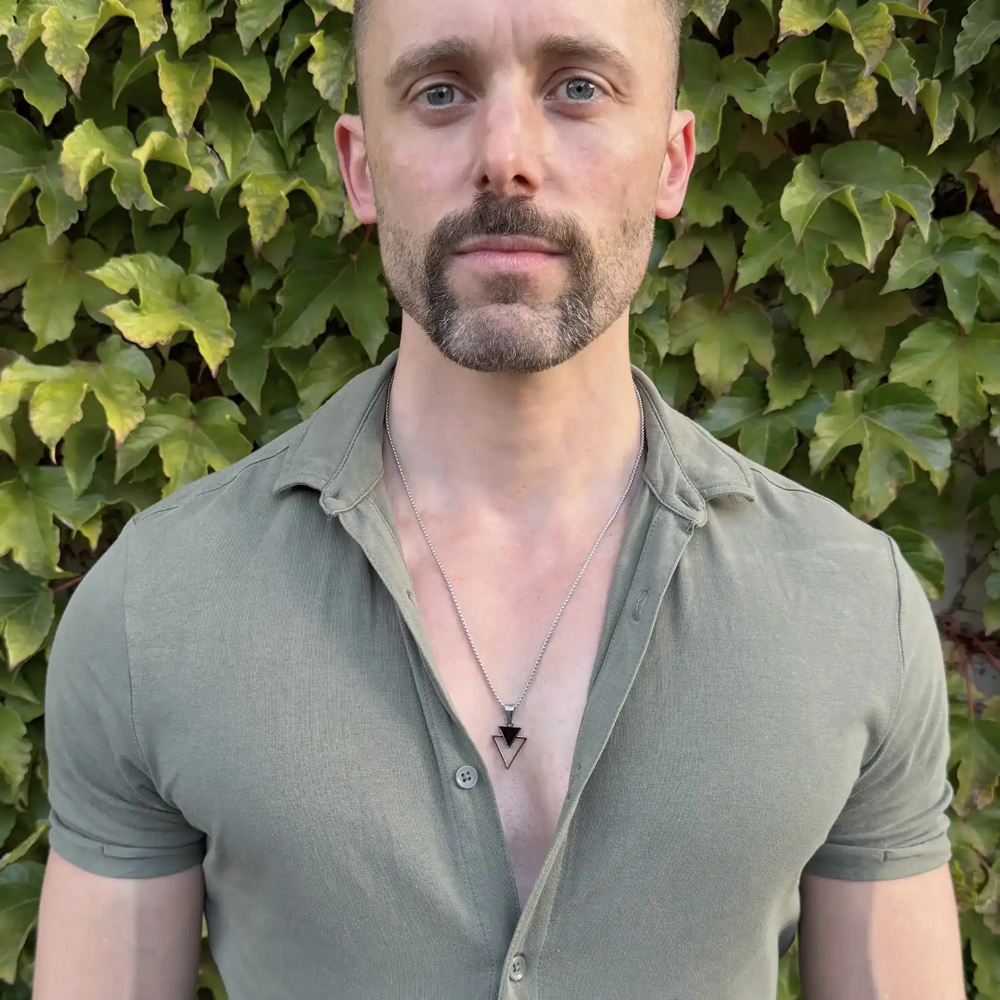 Bearded man standing against a green hedge wearing a stainless steel pendant necklace with a small black triangle stacked over an open triangle and a casual green button shirt