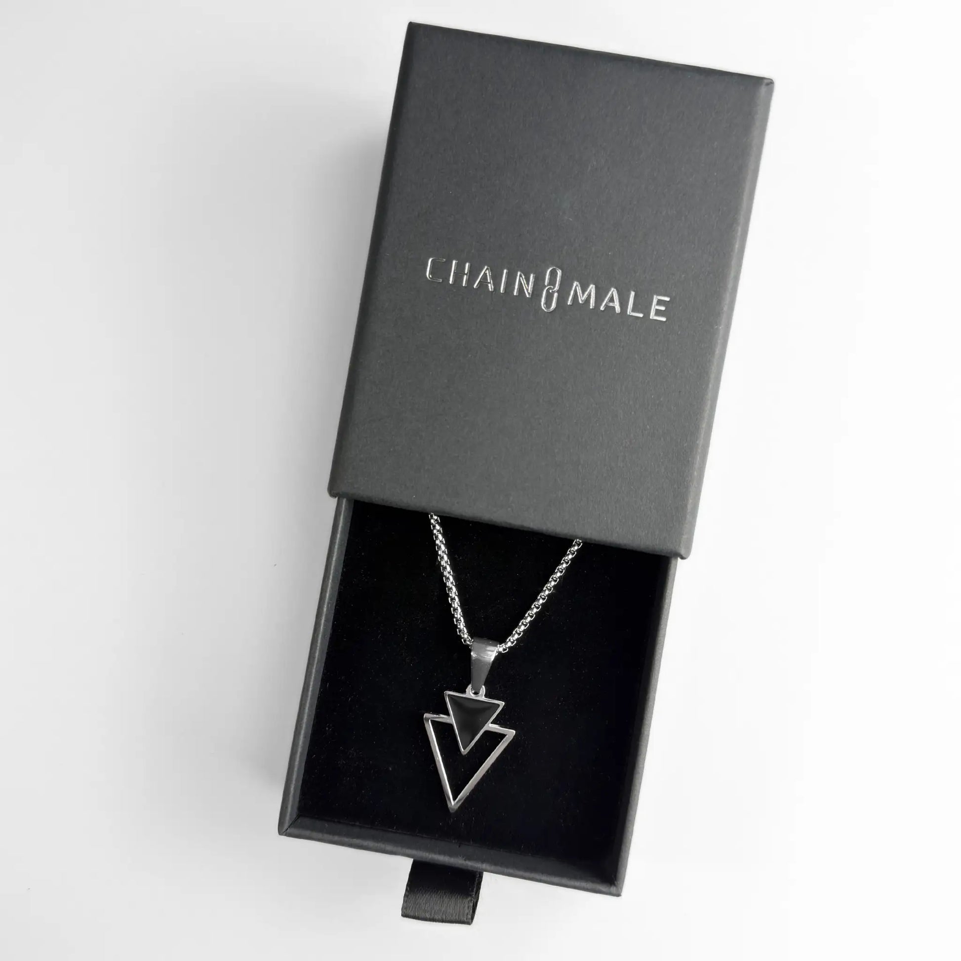 Stainless steel pendant necklace with a small black triangle stacked over an open triangle in a black Chain Male jewellery box on a white background