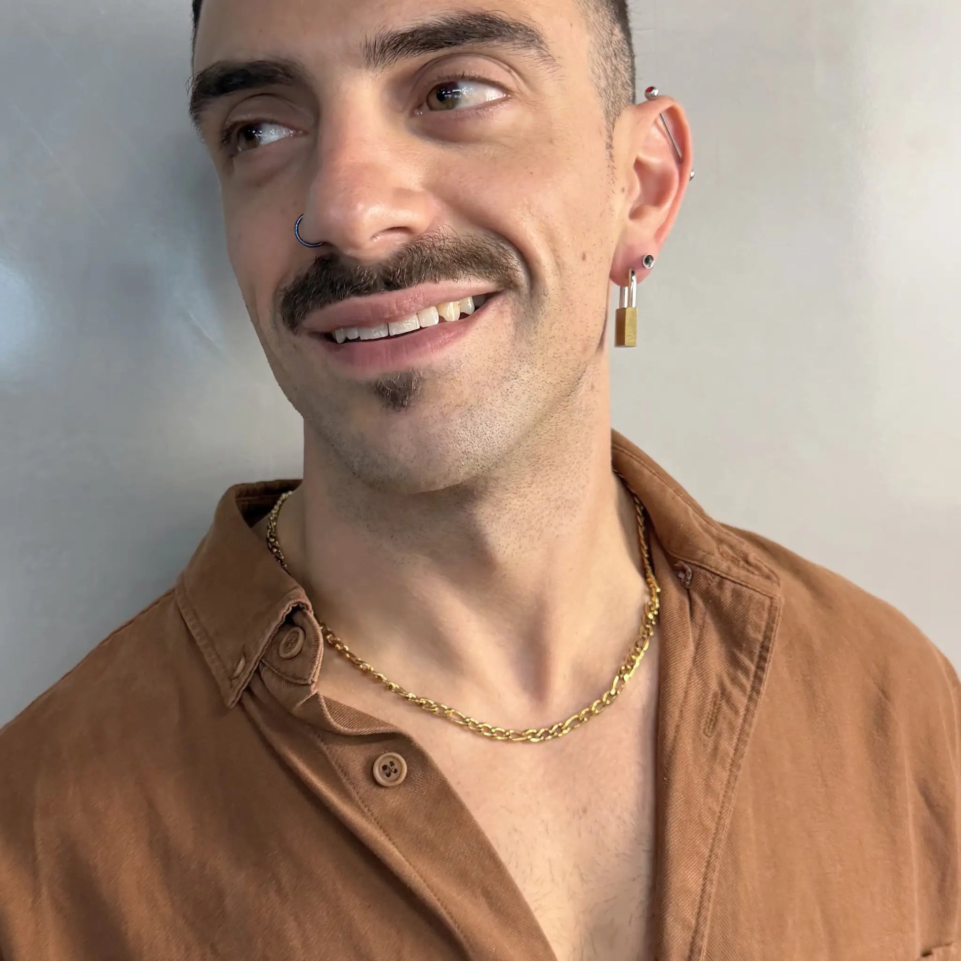 Man wearing a gold 5mm figaro chain necklace smiling in a brown shirt with piercings
