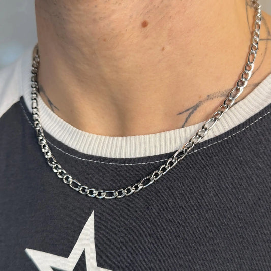 Close-up of a silver 5mm figaro chain necklace layered on a tattooed chest over a star print tee