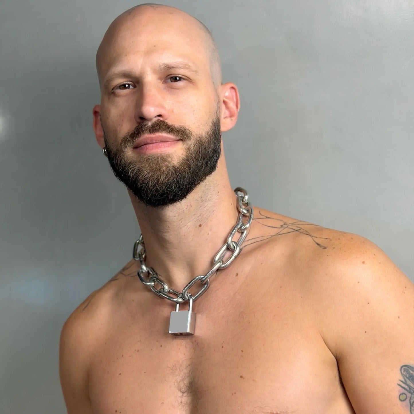 Tattooed bald man wearing a heavy duty stainless steel padlock chain necklace on a bare chest, facing camera