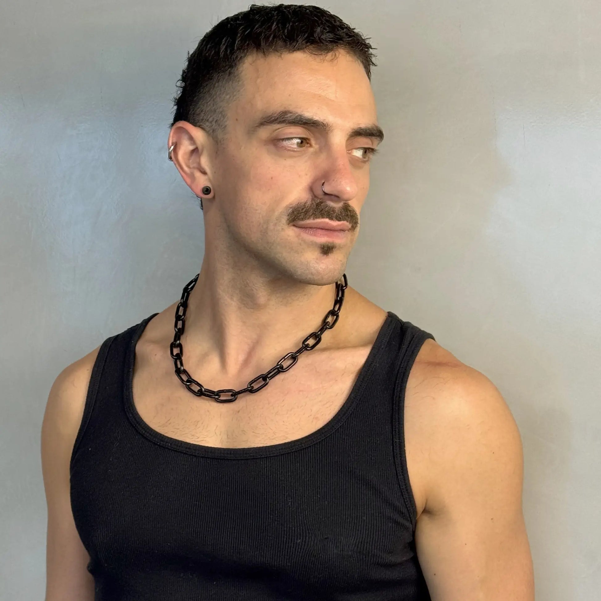 Man wearing a midnight black chain necklace in a black singlet, looking sideways with piercings and moustache