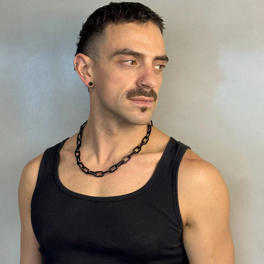 Man wearing a midnight black chain necklace in a black singlet, looking sideways with piercings and moustache