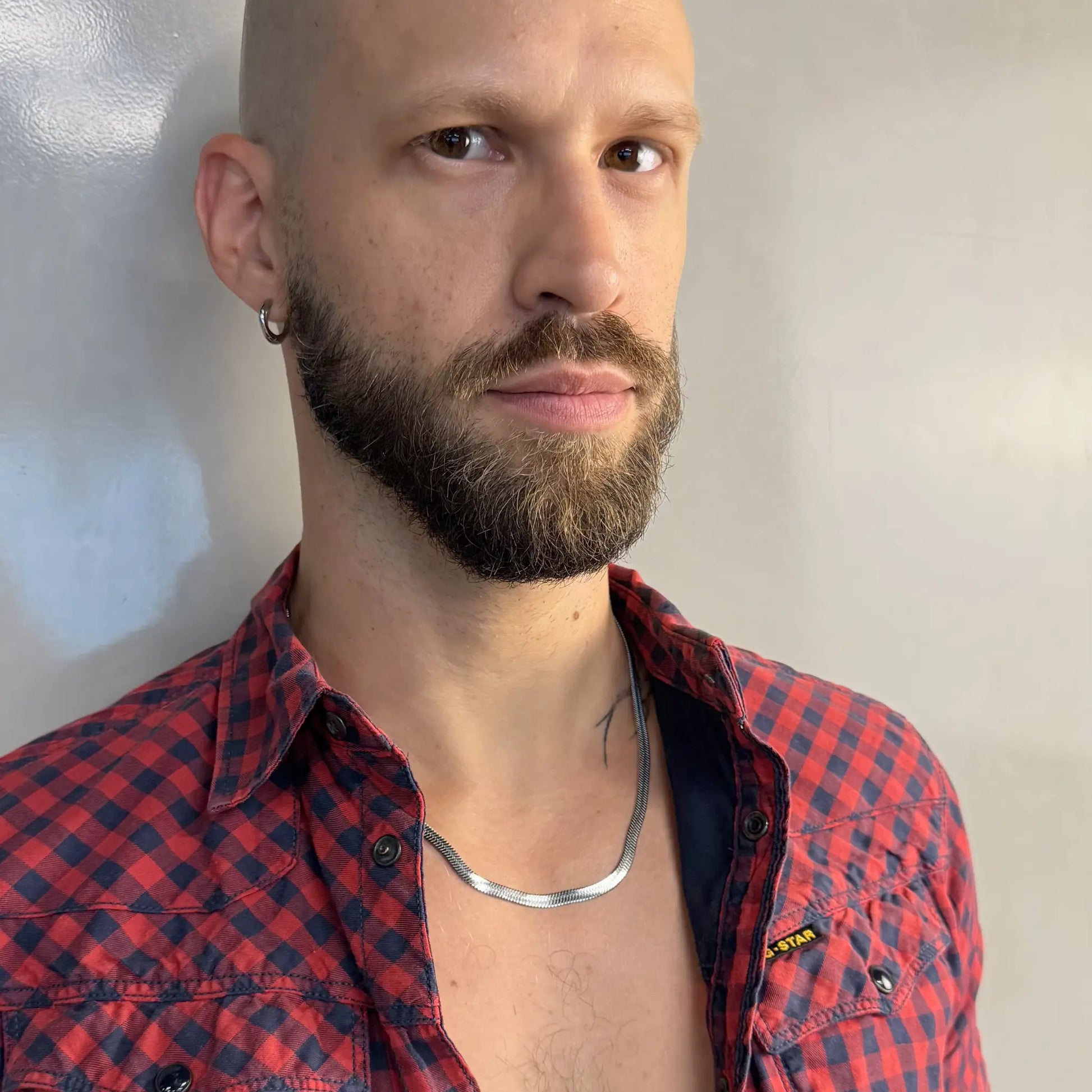 Tattooed bald man wearing a silver 5mm snake chain necklace in an open red flannel shirt facing camera