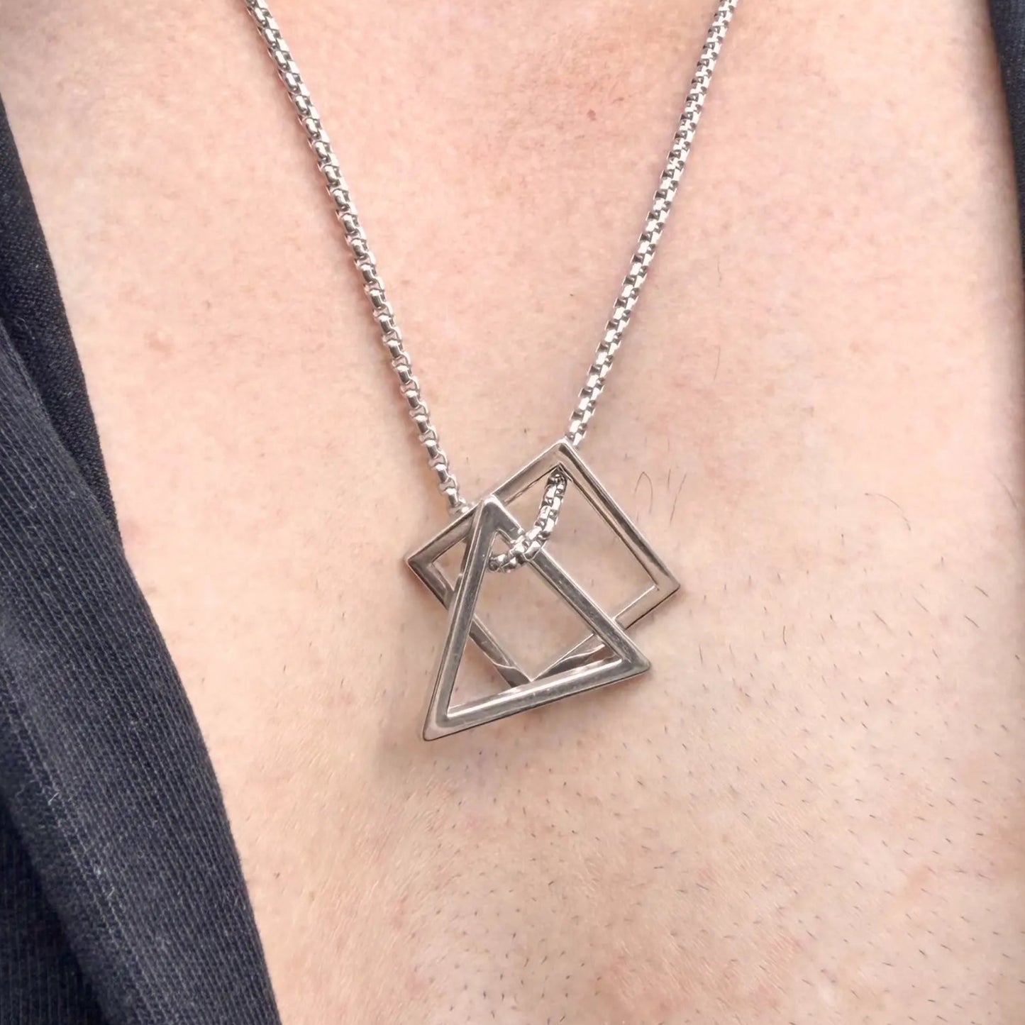 Close up of a stainless steel pendant necklace with interlocking square and triangle worn by a man with a black button shirt