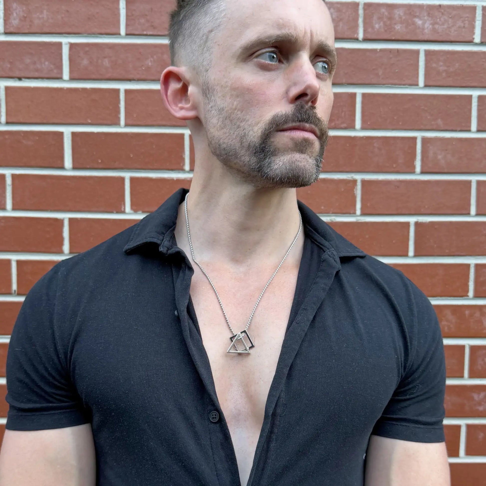 Stainless steel pendant necklace with interlocking square and triangle worn by a bearded man with a black button shirt against a terracotta brick wall