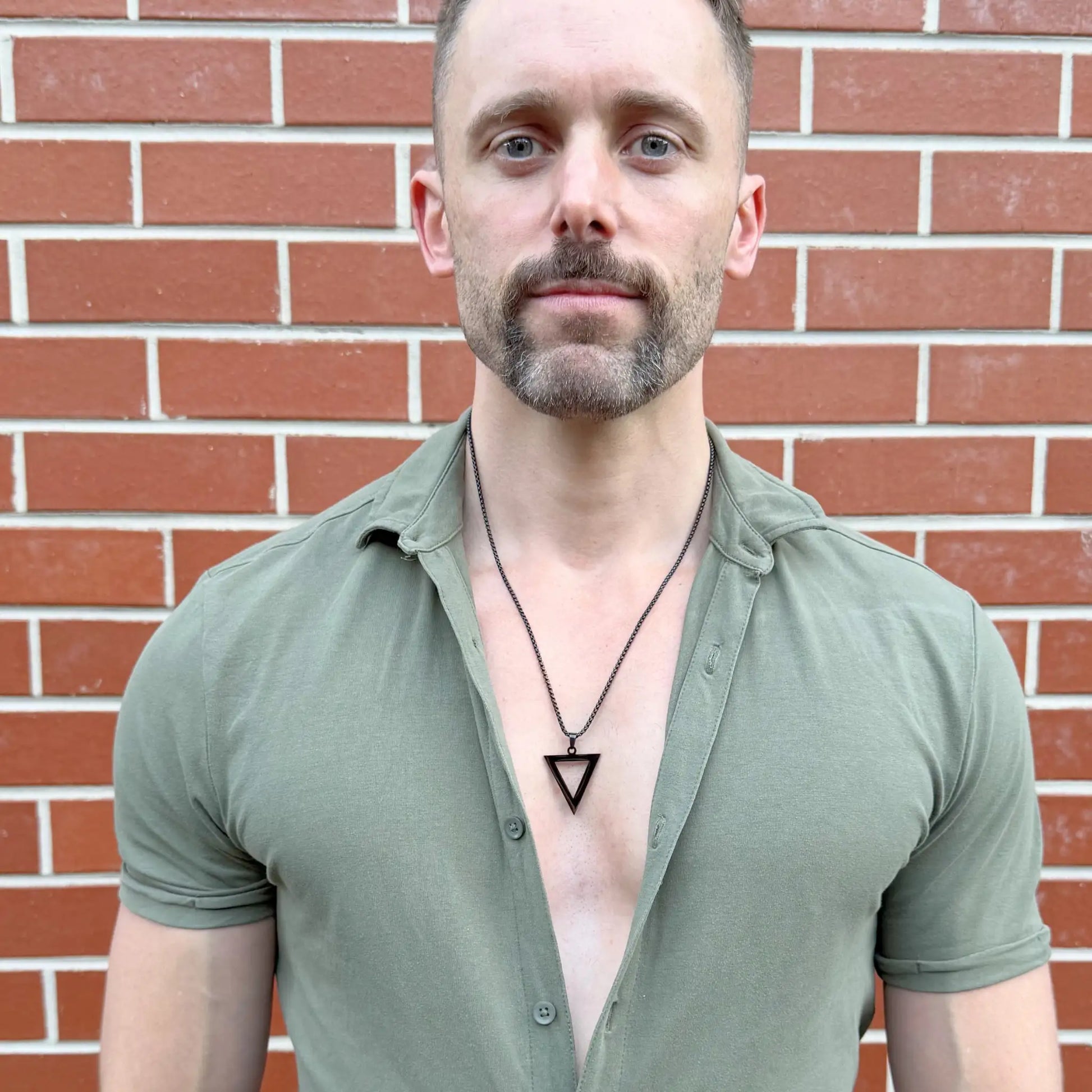 Bearded man with blue eyes wearing a black pendant necklace with a green button shirt against a terracotta coloured brick wall