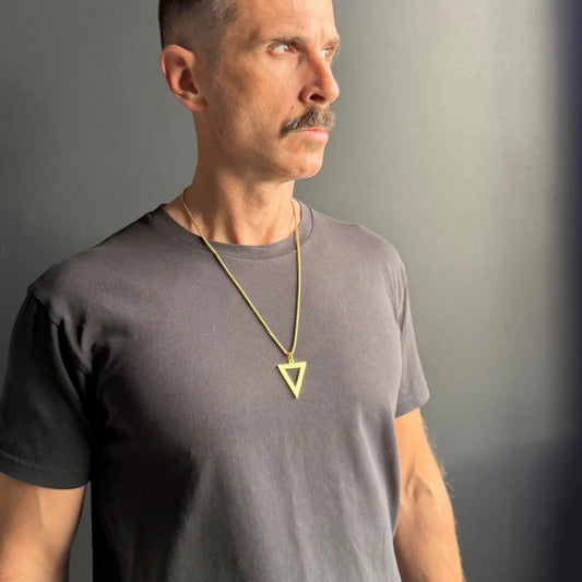Gold triangle pendant necklace worn by a man with a moustache wearing a black tshirt against a dark grey background