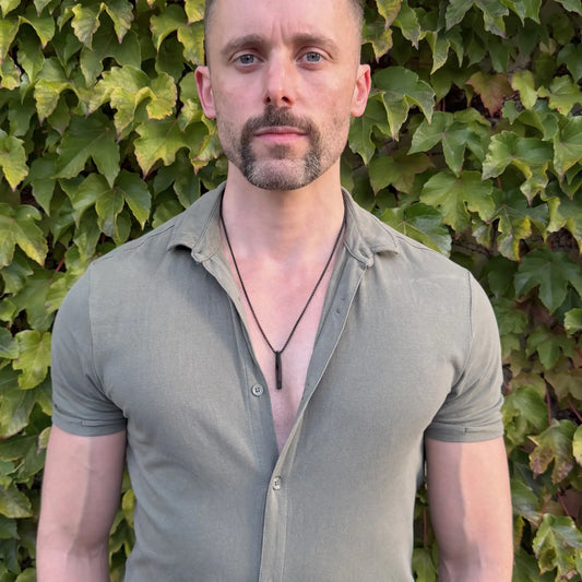 Bearded man wearing a black bar pendant necklace and a green button shirt in front of a thick green hedge