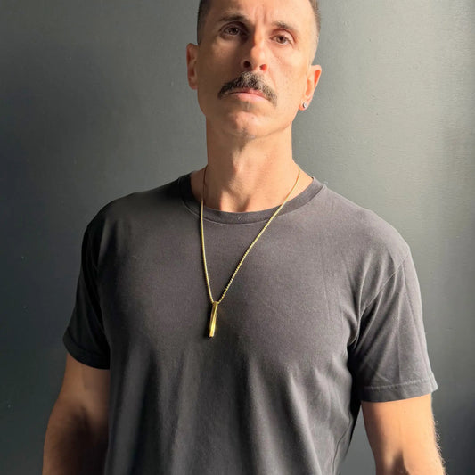 Man with moustache wearing a gold plated stainless steel bar pendant necklace over a black tshirt against a dark grey wall