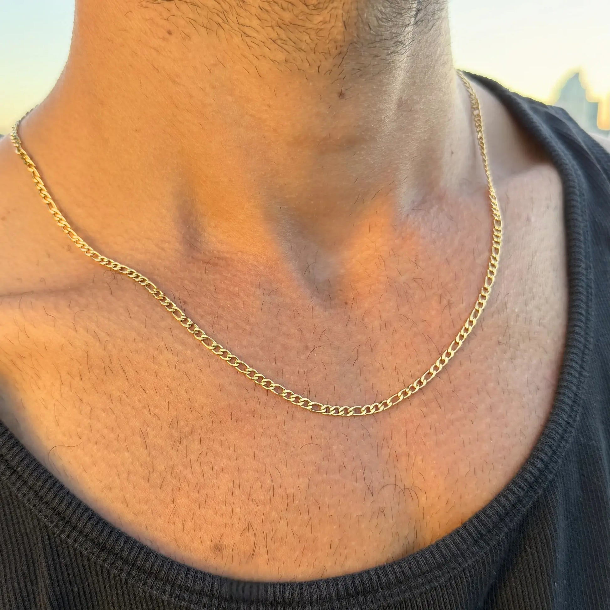 Gold 3mm figaro chain necklace worn by a man in a black singlet at sunset