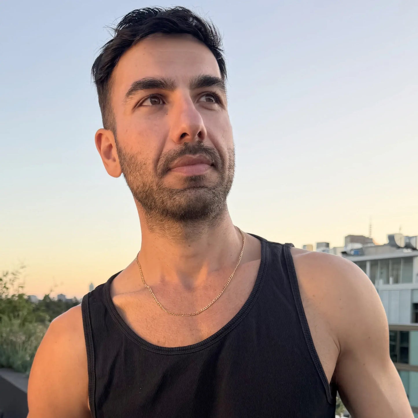 Man wearing a gold figaro chain necklace outdoors at golden hour with city skyline in background