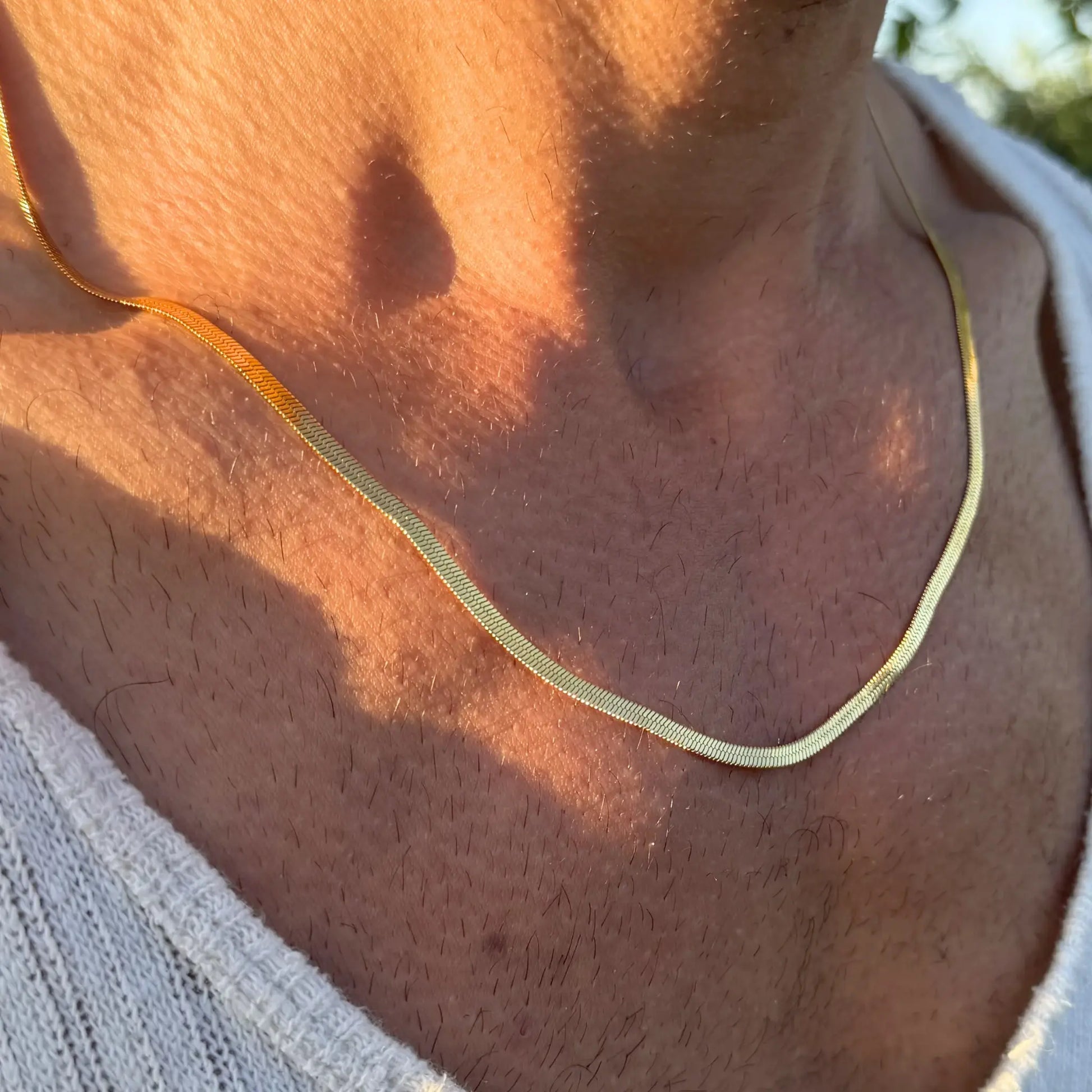 Close-up of a gold 3mm snake chain necklace worn with a white singlet at golden hour with dappled light