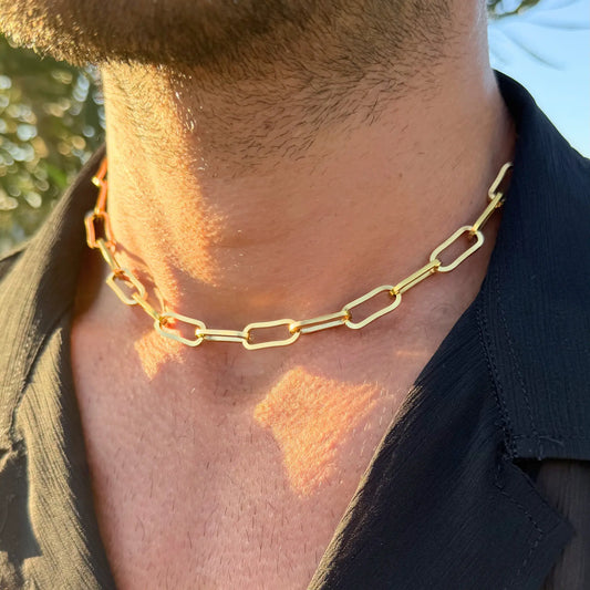Close-up of a gold paperclip choker necklace worn by a man in a dark shirt outdoors at golden hour