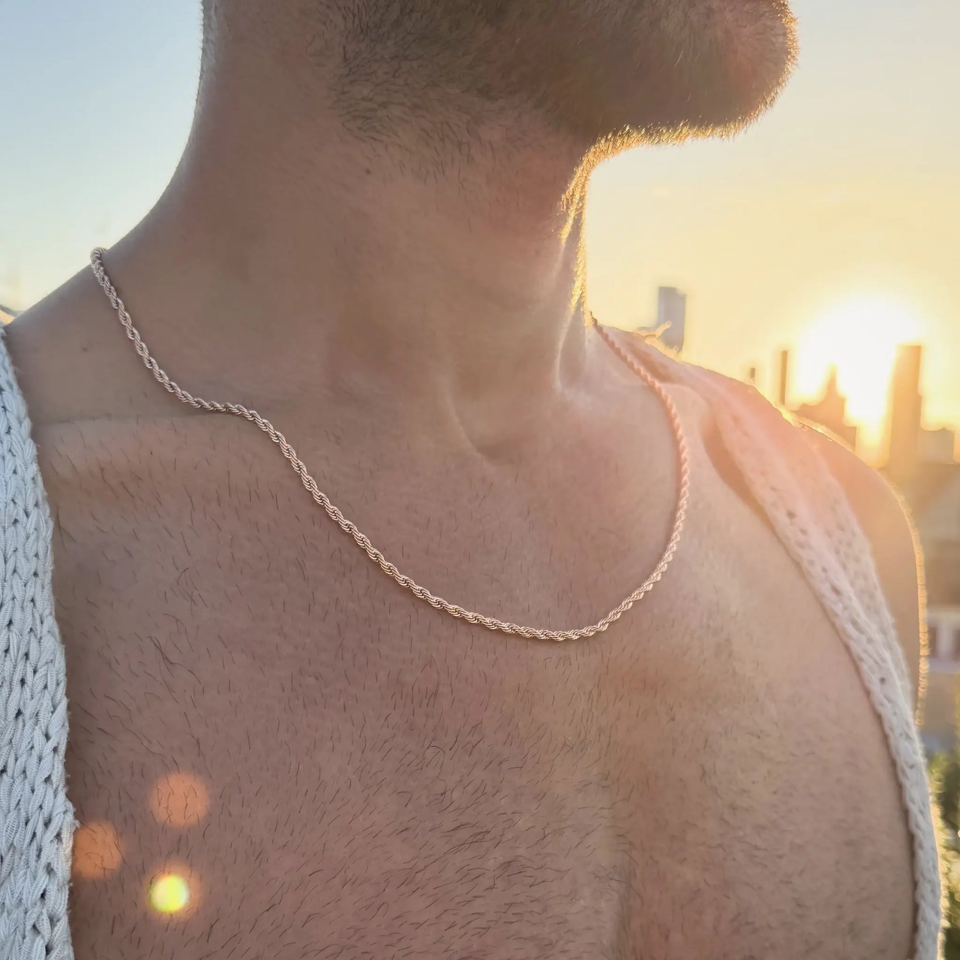 Man wearing a rose gold rope chain necklace in a white knit vest outdoors at sunset