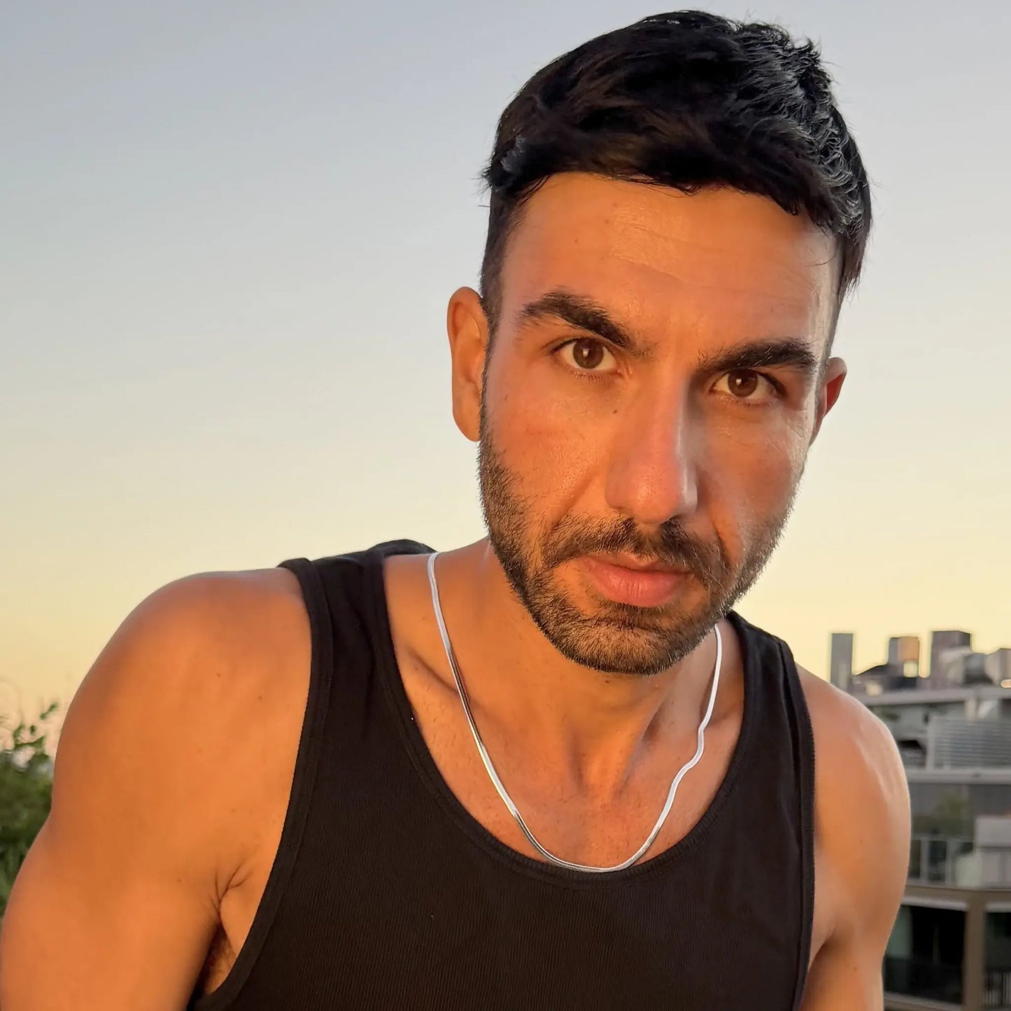 Man wearing a silver 3mm snake chain necklace looking directly at camera with city skyline at dusk